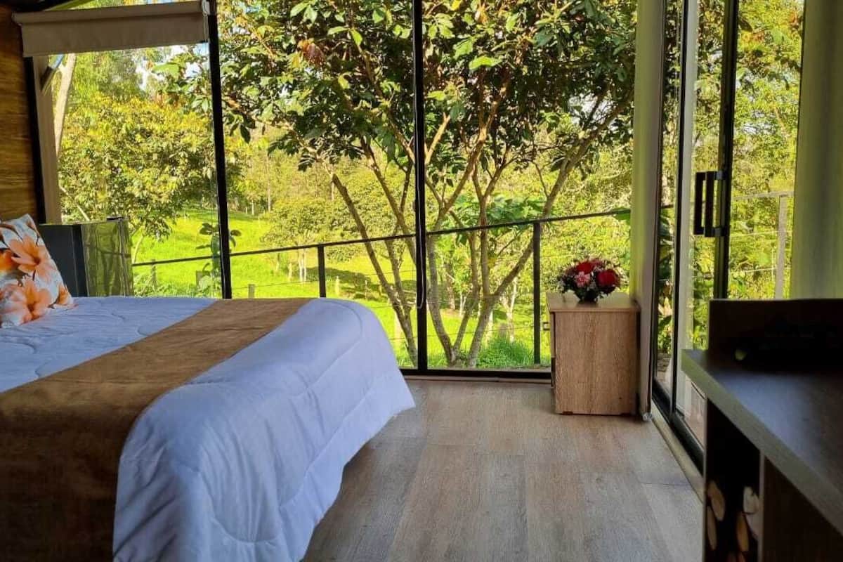 A bedroom with a large bed, a wooden headboard, a nightstand, and a window with a view of greenery.