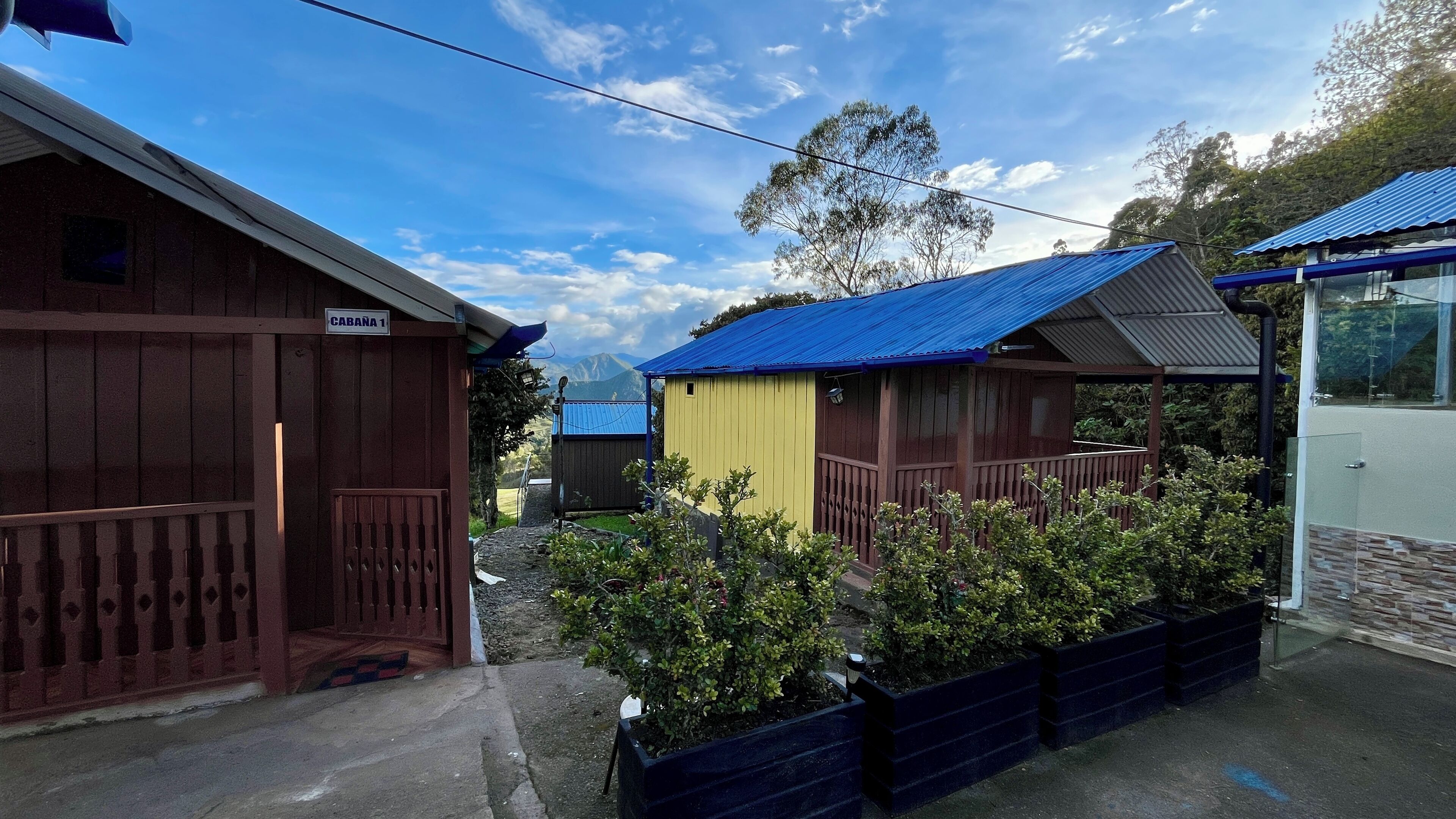A row of cabins with wooden exteriors and blue roofs, surrounded by greenery and a clear sky.