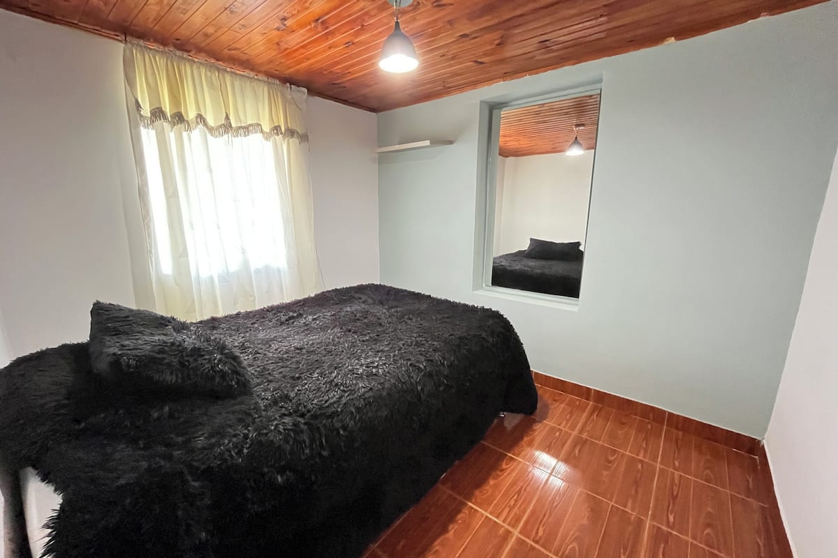 A bedroom with a wooden ceiling, a bed with a dark blanket, a window with blinds, and a mirror on the wall.