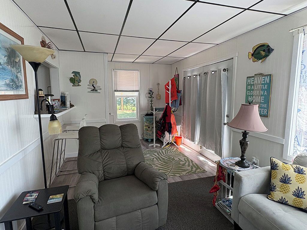 Sala de estar com sofá, poltrona e luminária. Há uma janela com cortinas, uma porta e uma estante com objetos.