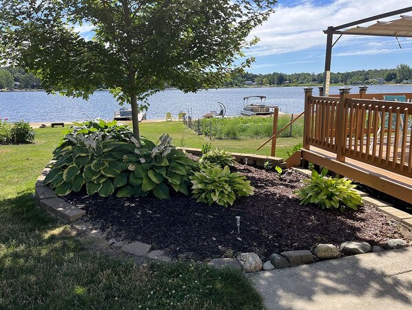 A lakeside deck with a wooden railing, a stone pathway, and a variety of plants.