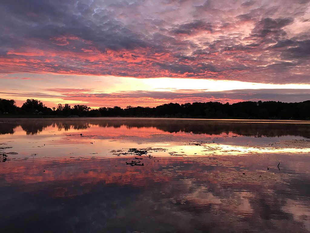 Un lac paisible au coucher du soleil, dont la surface reflète le ciel, et qui est entouré d’arbres.