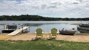 Une jetée au bord du lac, avec deux chaises Adirondack vertes et une petite table, ainsi qu’un bateau et un autre bateau amarré.