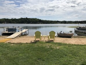 A lakeside dock with two green Adirondack chairs and a small table, a boat, and a docked boat.