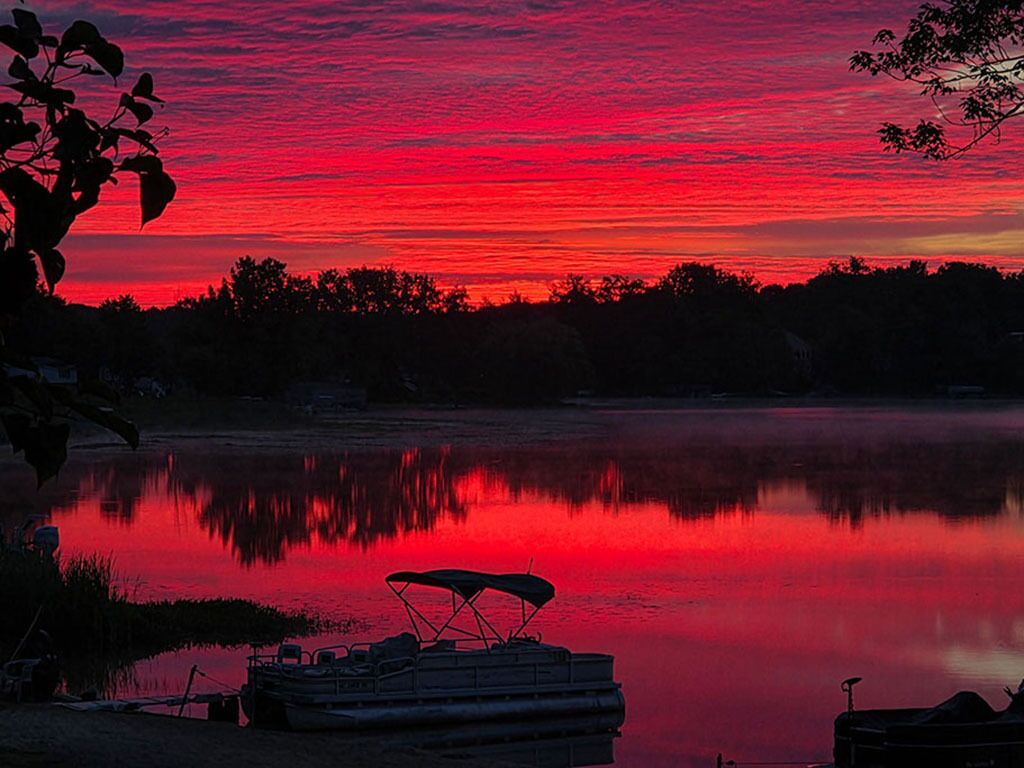 A calm lake at sunset with a red sky, a dock with a boat, and trees in the background.