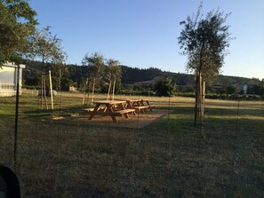 Un área de picnic con bancos de madera y un cerramiento, rodeada de árboles y un paisaje montañoso al fondo.