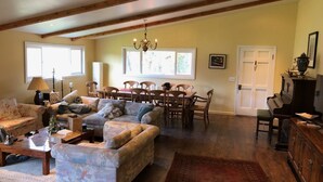 A spacious living room with a large sofa, a dining table with chairs, a chandelier, and a piano.