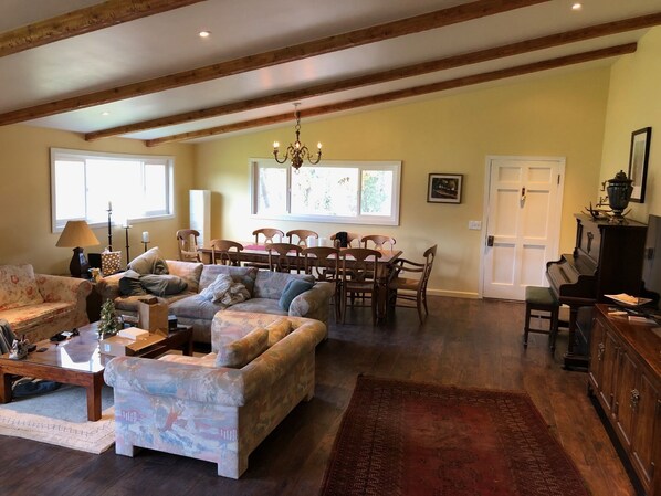 A spacious living room with a large sofa, a dining table with chairs, a chandelier, and a piano.
