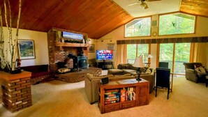 A cozy living room with a fireplace, comfortable seating, a television, and large windows offering a view of the outdoors.