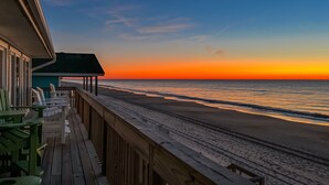 Een houten terras met stoelen en tafels met uitzicht op een strand bij zonsondergang.