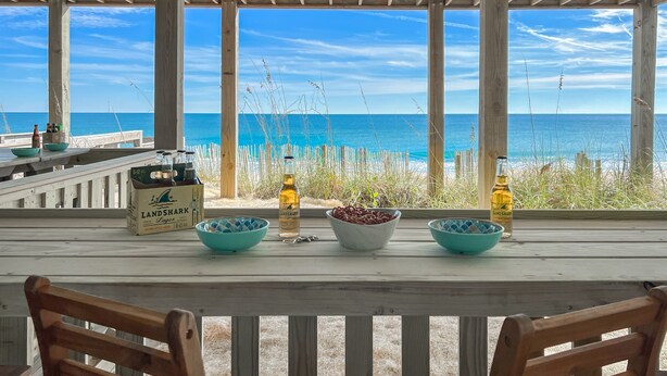 Een houten terras met uitzicht op de oceaan, een tafel met drankjes en snacks, en een stoel.