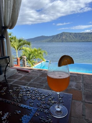 A glass of amber liquid with a garnish, a jigsaw puzzle, and a view of a pool and mountains.