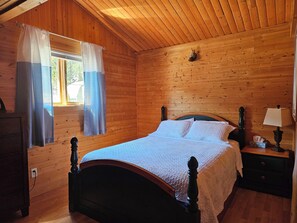 Un dormitorio de cabaña de madera con cama, cortinas, una mesita de noche y una lámpara.