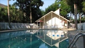 Une piscine aux eaux cristallines, bordée de chaises longues et d’ombrelles, et entourée d’une clôture et d’arbres.