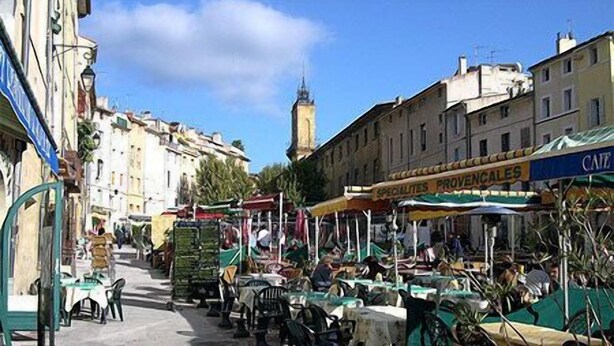 Un café en plein air, avec des tables et des chaises, entouré d’immeubles et d’un beffroi.