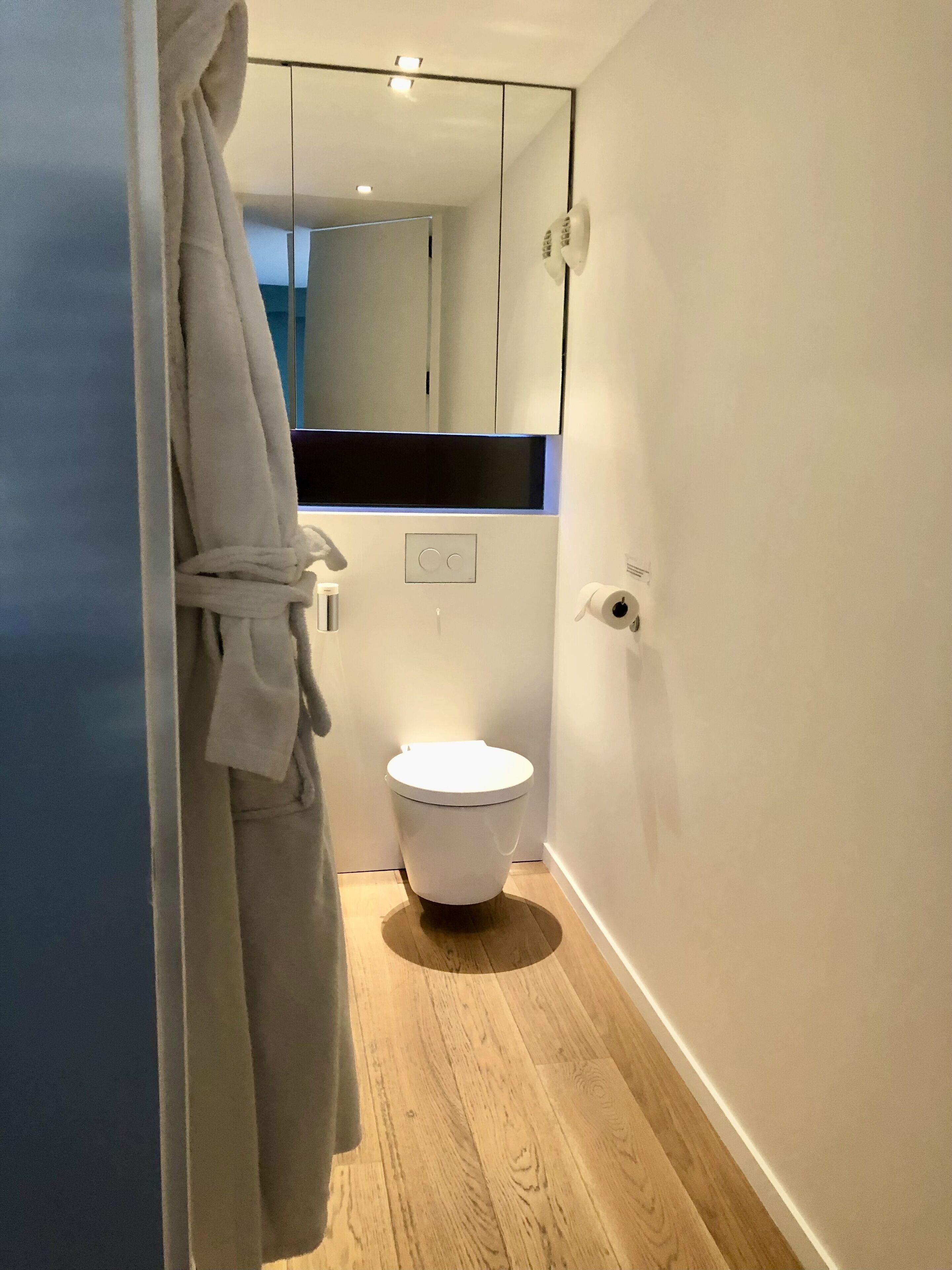 Un baño moderno con inodoro blanco, suelo de madera y una bata colgada en la puerta.