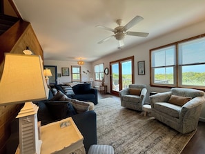 A spacious living room with a ceiling fan, large windows, and a view of the outdoors.