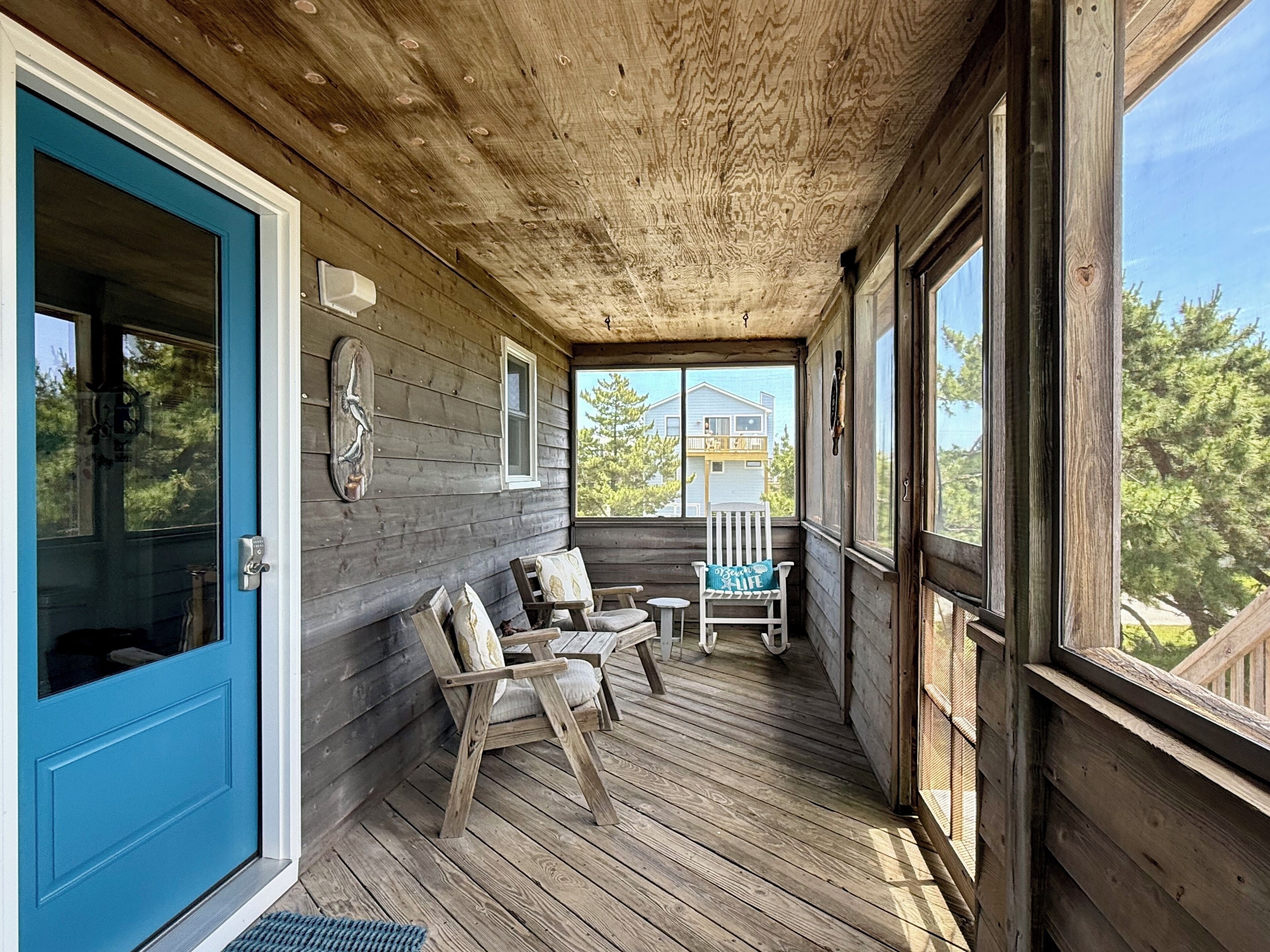 A wooden porch with a blue door, two chairs, a small table, and a view of a house in the background.