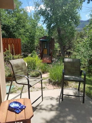 Un patio con due sedie e un tavolo, una piccola struttura simile a un santuario sullo sfondo, circondato da vegetazione e un recinto in legno.