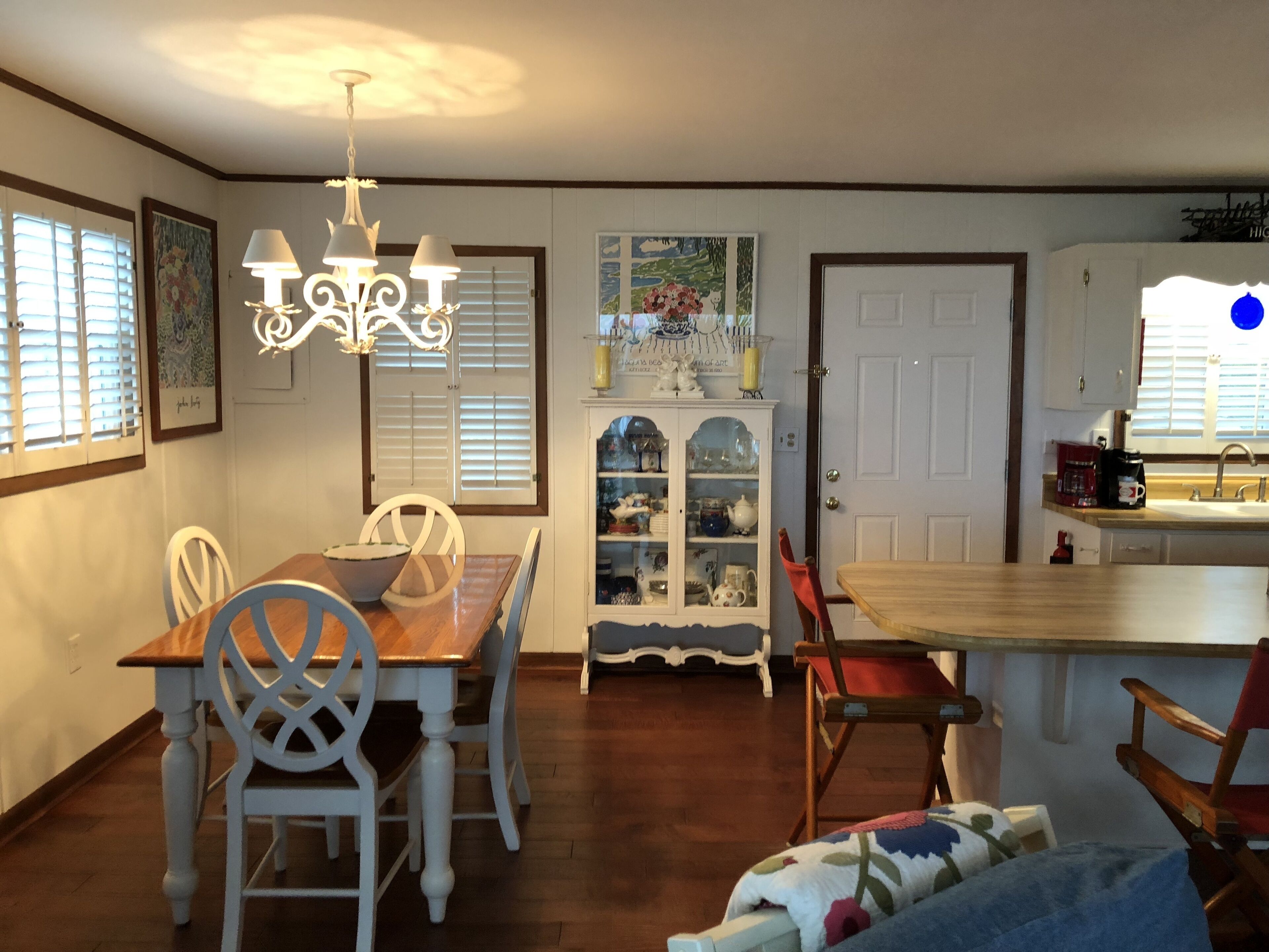 A dining area with a wooden table and white chairs, a chandelier, a cabinet with glass doors, and a kitchen area with a sink and coffee maker.