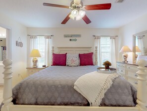 A bedroom with a bed, a ceiling fan, a nightstand with a lamp, a dresser with a mirror, and a window with curtains.