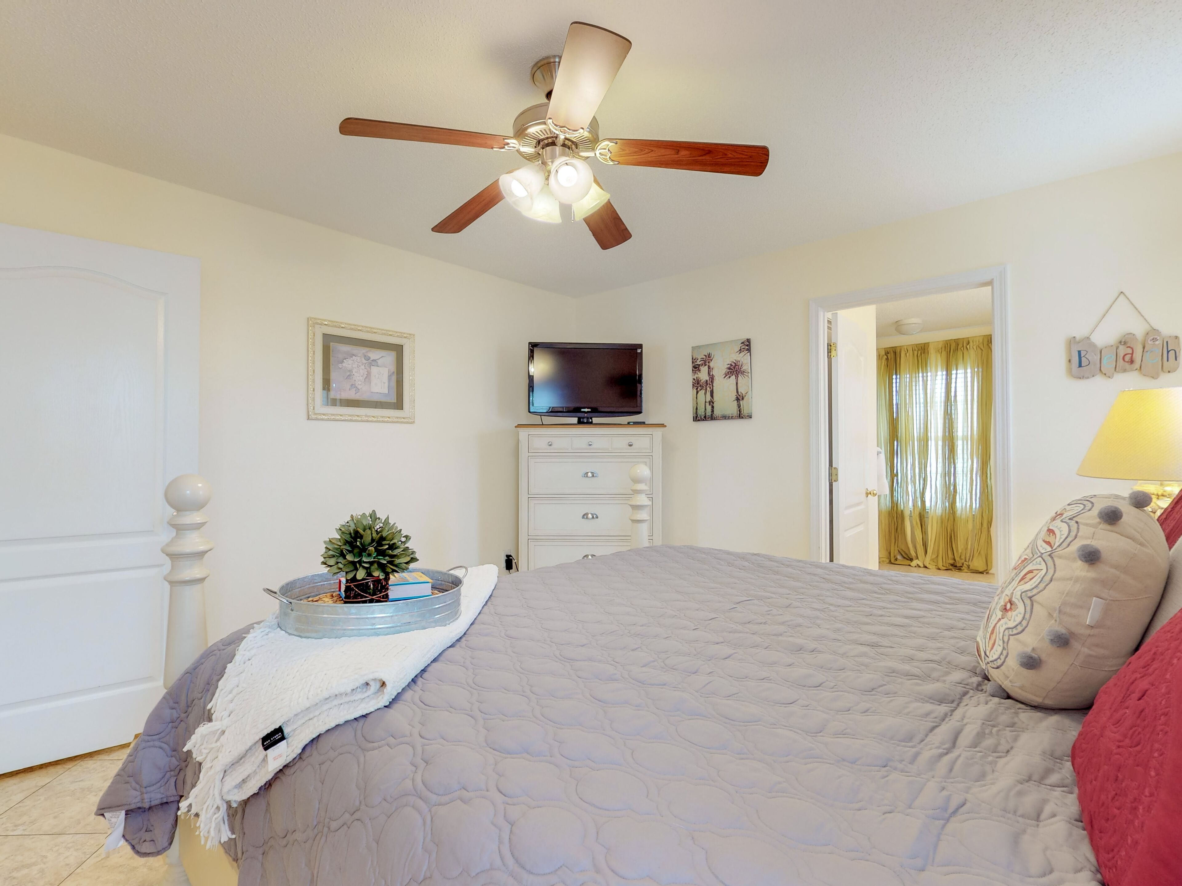 Une chambre à coucher comprenant un lit, un ventilateur de plafond, un téléviseur, une commode, une plante et une porte donnant sur une autre pièce.