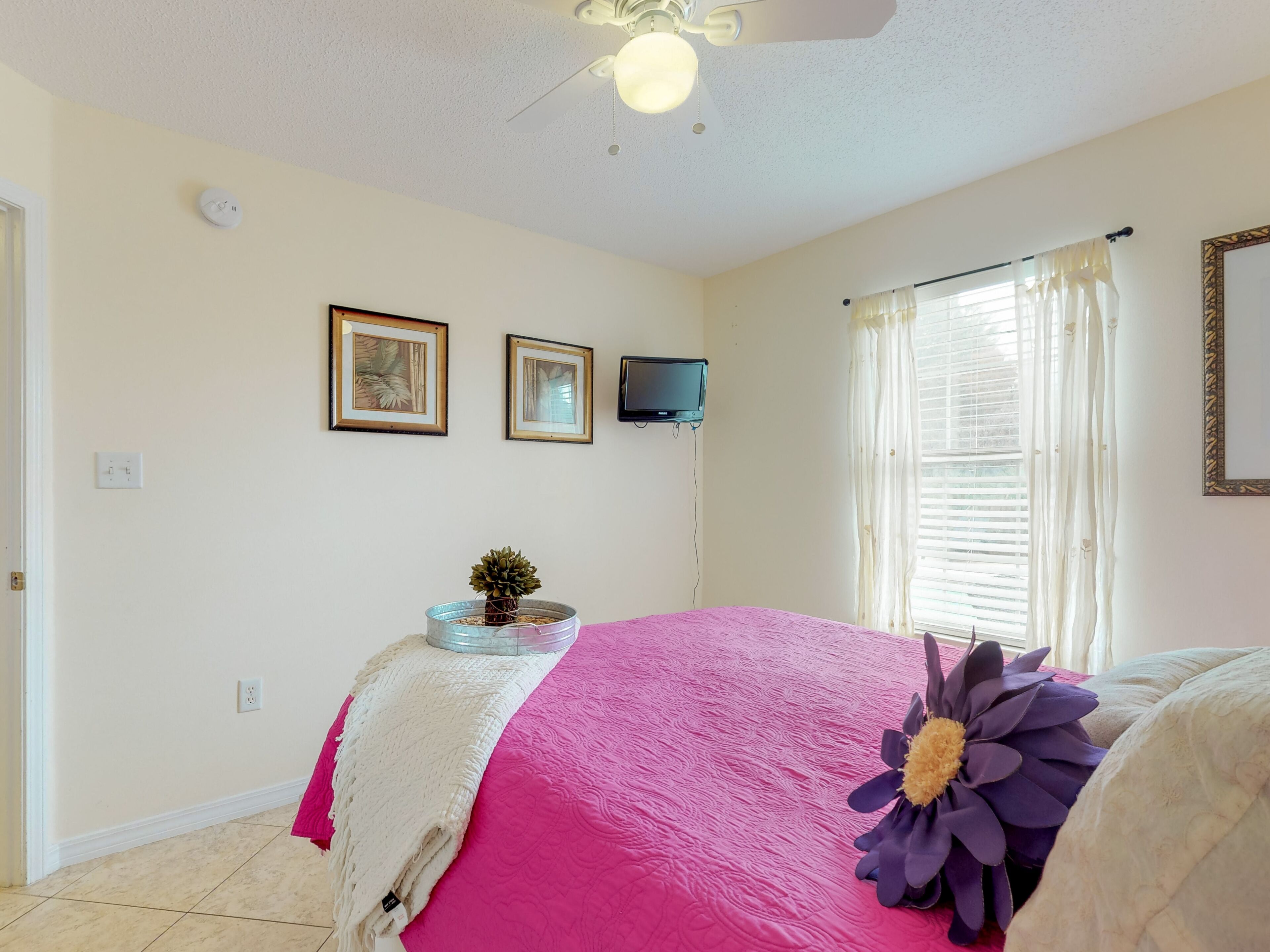 A bedroom with a bed, a ceiling fan, a wall-mounted TV, and a window with curtains.