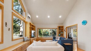 A spacious living room with a large window, built-in shelving, and a cozy seating area. - Family Favorite! Treehouse,Huge Yard,Firepit & EV Charger.Walk to town & beach (Nehalem)