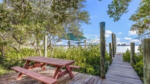 A wooden deck with a picnic table, surrounded by greenery and leading to a dock over water.