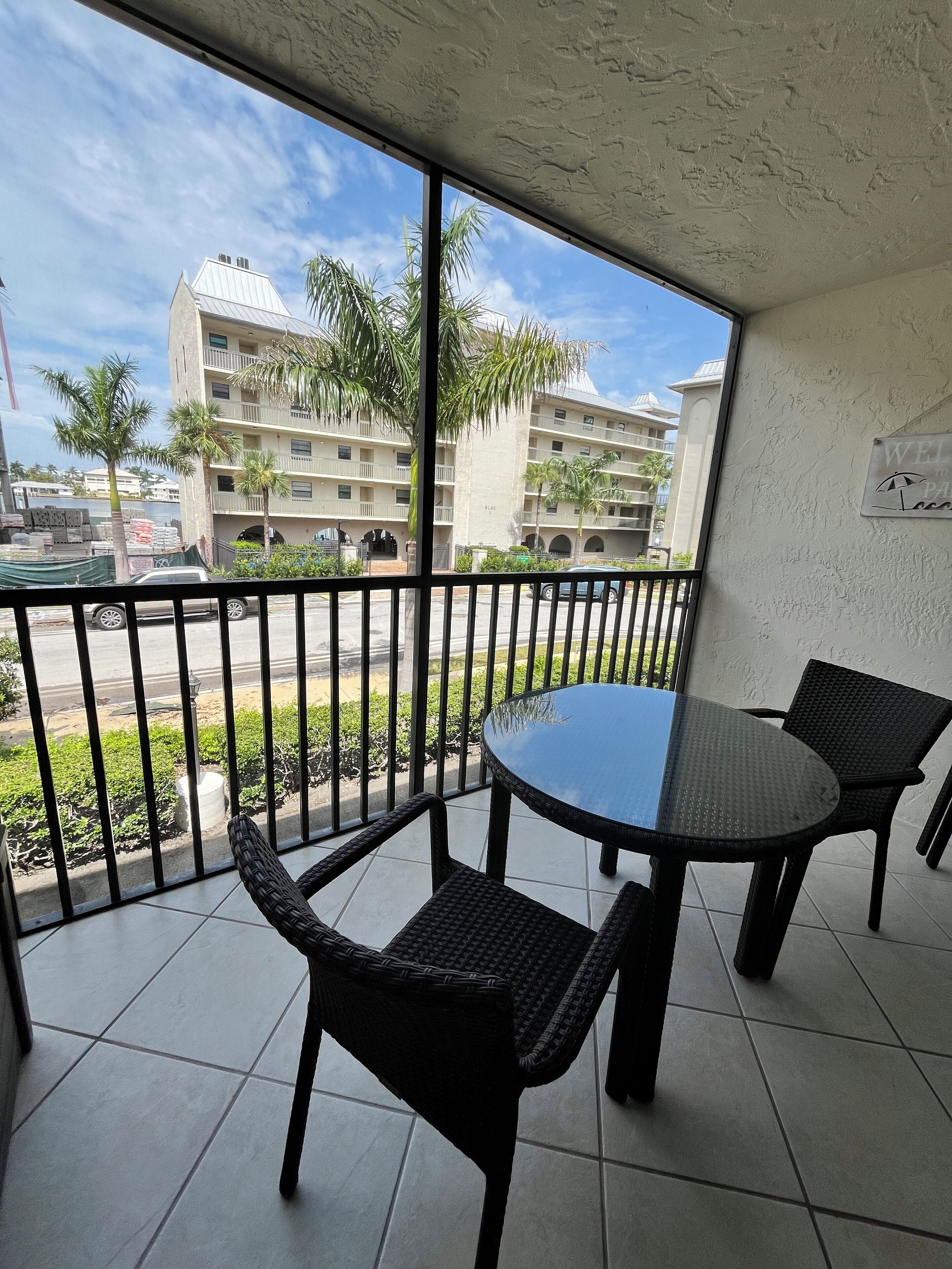 Een balkon met een ronde tafel en twee rieten stoelen, met uitzicht op een gebouw met meerdere balkons en palmbomen.