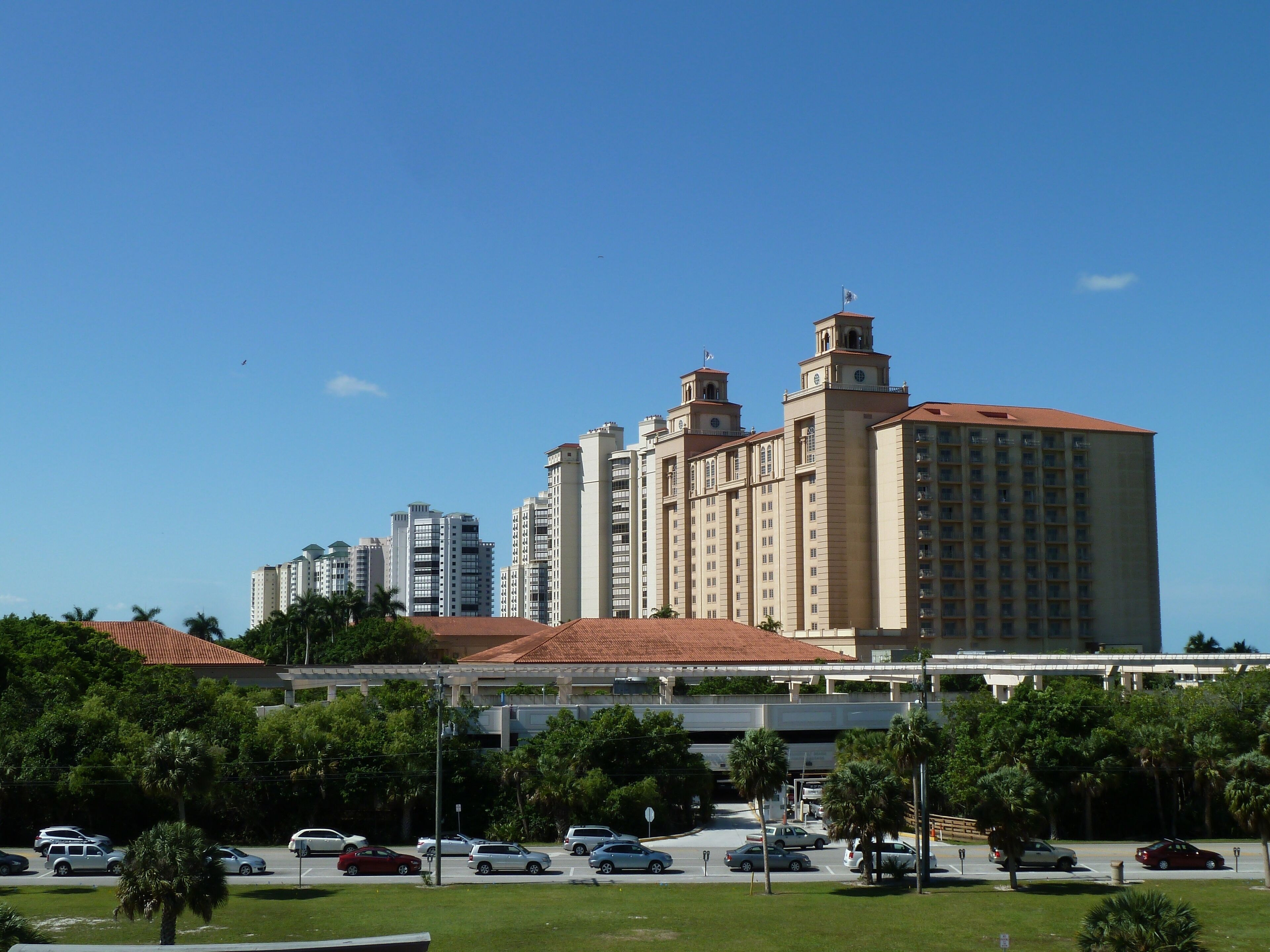 A large hotel with multiple towers, a parking lot with cars, and a clear blue sky.