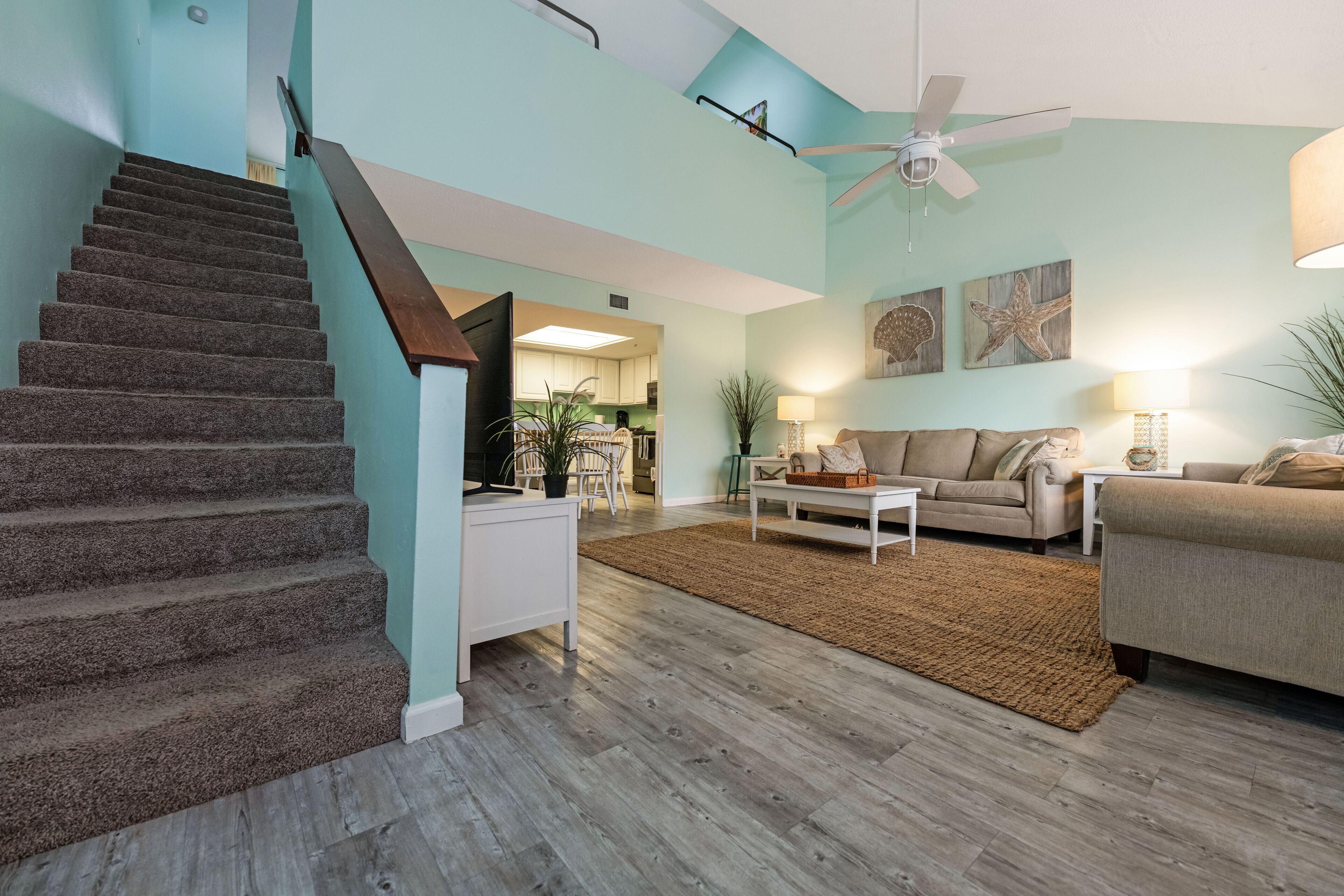 A spacious living area with a staircase, a ceiling fan, and a kitchen in the background.