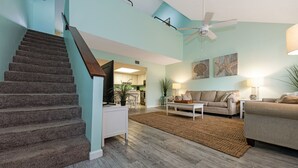 A spacious living area with a staircase, a ceiling fan, and a kitchen in the background.