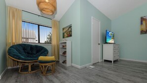 A room with a large window, a wicker chair, a television on a cabinet, and a bookshelf.