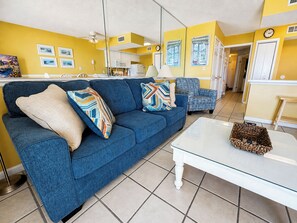 Una sala de estar con un sofá azul, una mesa de centro de vidrio y una cocina al fondo.