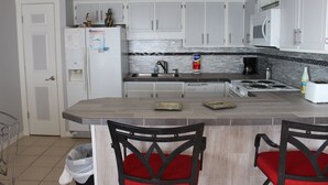 Cocina con armarios blancos, salpicadero azulejado, estufa y lavamanos. Hay dos sillas con cojines rojos y un cubo de basura.