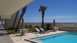 Zona de piscina con sillas de descanso y sombrillas, con vista despejada al océano y una valla blanca.