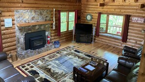 A log cabin interior with a stone fireplace, leather seating, a television, and a view of the outdoors.