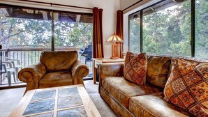 Ein Wohnzimmer mit Ledersofas, einem Couchtisch und einer Lampe. Große Fenster mit Vorhängen bieten Blick auf Bäume.
