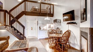 Ein geräumiges Wohnzimmer mit einer Holztreppe, einem Essbereich mit Holzbänken, einem an der Wand montierten Fernseher und einem Kamin.