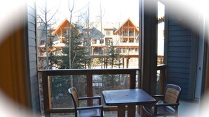 A balcony with a table and two chairs, overlooking a building with multiple windows and a view of trees. - Enjoy Mountain Views @ Fully Equipped 2BR2BA Hotel Condo (Canmore)