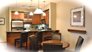 A modern kitchen with wooden cabinets, a stainless steel refrigerator, a built-in microwave, and a granite countertop. There are four black chairs around a wooden dining table. - Enjoy Mountain Views @ Fully Equipped 2BR2BA Hotel Condo (Canmore)