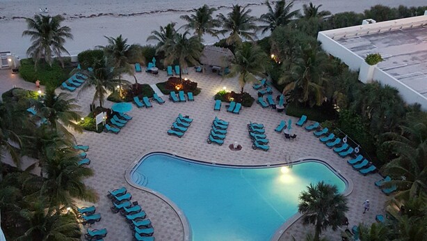 Una piscina di un resort con lettini disposti a cerchio, circondata da palme, con vista sulla spiaggia e sull'oceano sullo sfondo.
