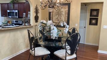 A dining area with a round table, four chairs, a chandelier, and a kitchen in the background.