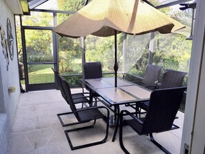 Um alpendre coberto com mesa e cadeiras, janelas amplas e vista para o jardim.