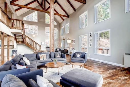 A spacious living room with a high ceiling, large windows, and a staircase leading to an upper level.