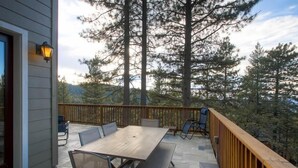 Un patio exterior con una mesa y sillas de madera, barandilla de madera y vistas a árboles altos.