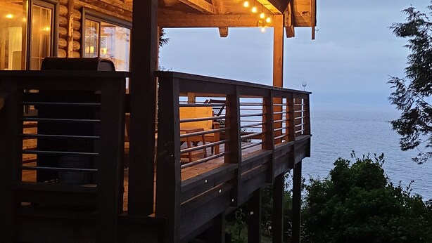 A wooden cabin with a balcony overlooking a body of water, string lights, and a grassy area with daisies. - An Oceanfront Cottage with Stunning Views and Direct Beach Access (Shirley)