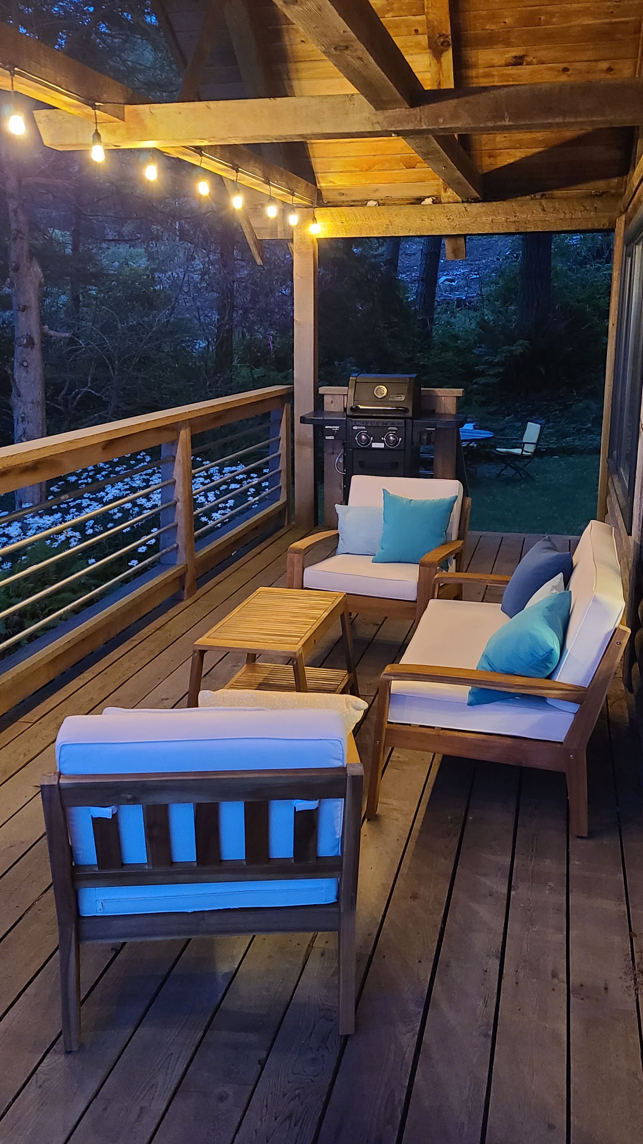 Terraza exterior con mobiliario de madera, una parrilla y luces colgantes.
