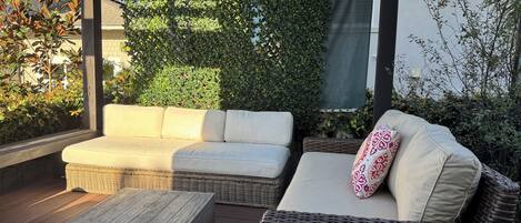 Un patio exterior con mobiliario de mimbre, incluyendo un sofá y un sillón de dos asientos, una mesa de centro de madera y una estructura de pérgola.